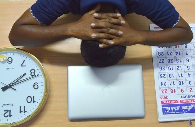  Een oververmoeide man houdt zijn hoofd op zijn bureau, naast zijn laptop, agenda en klok